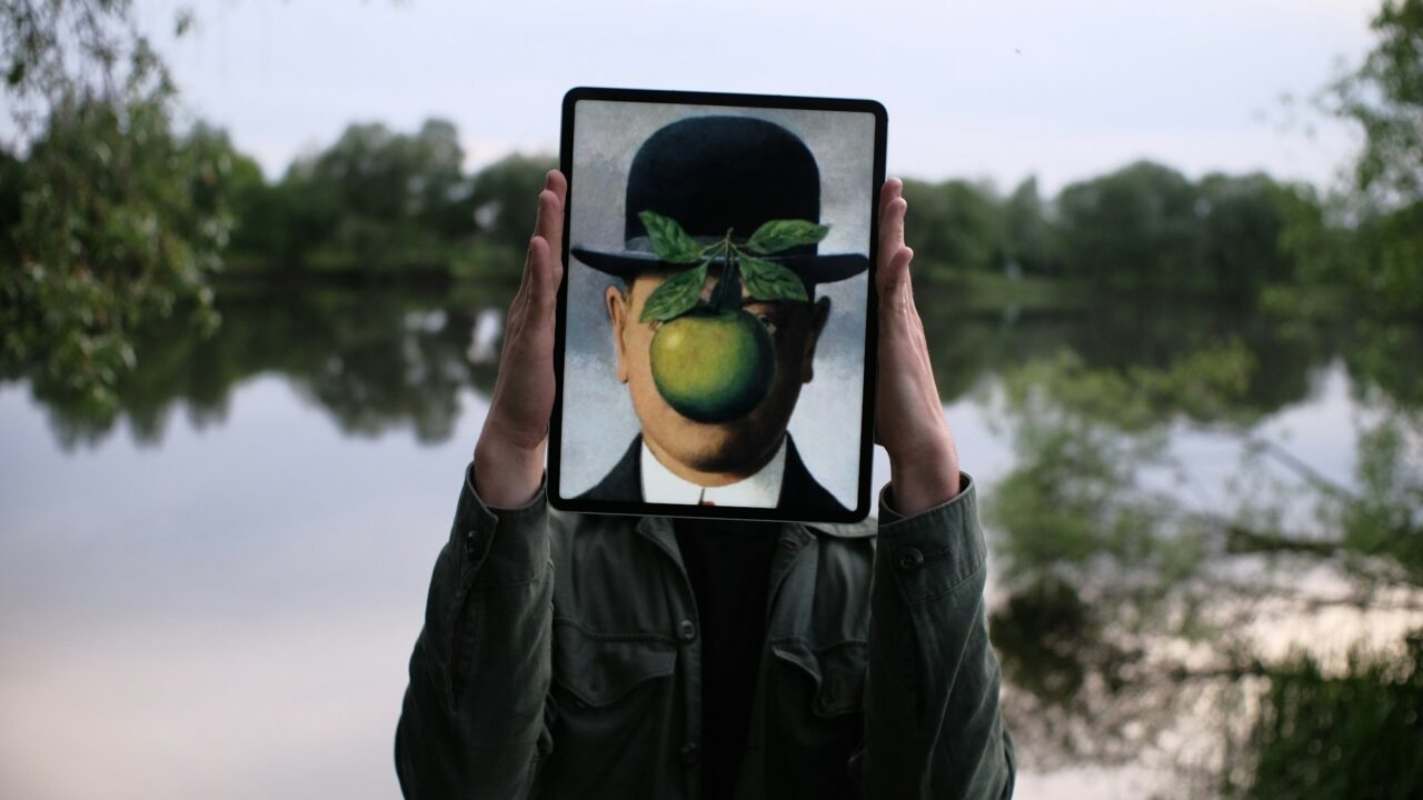 A person outdoors holds up a tablet showing an image of a face obscured by a bowler hat and green apple, the digital image covering their own face