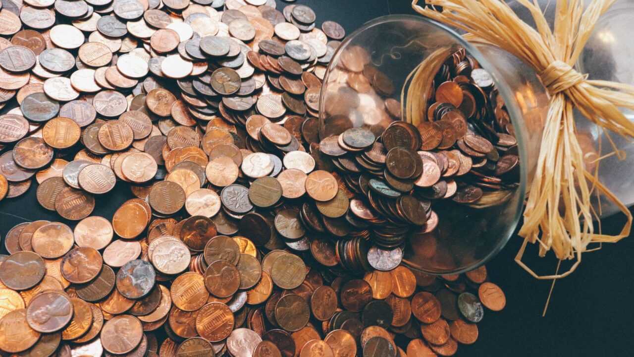 A clear glass jar on its side with copper coins scattered across a wooden surface.
