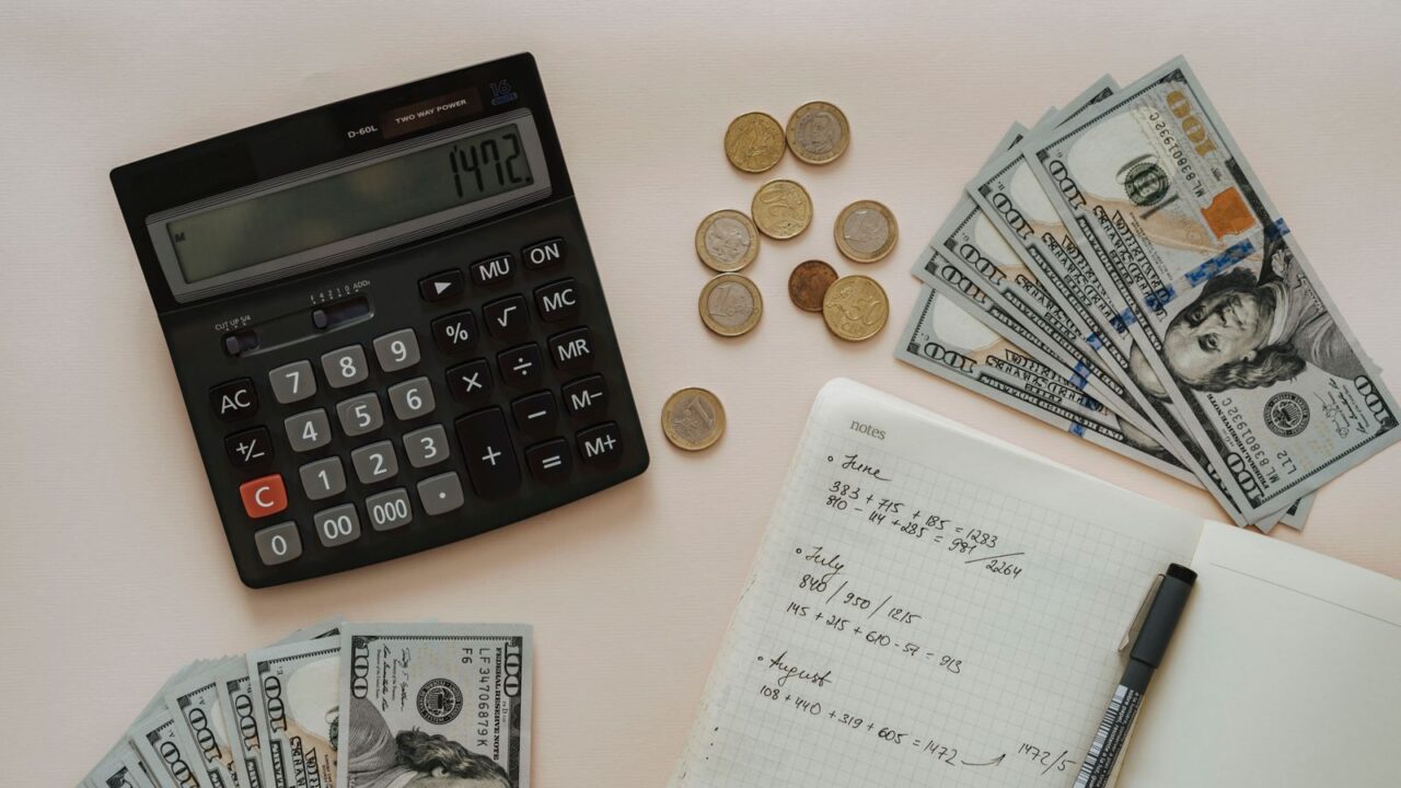 Calculator beside banknotes, coins and handwritten financial notes on a desk.