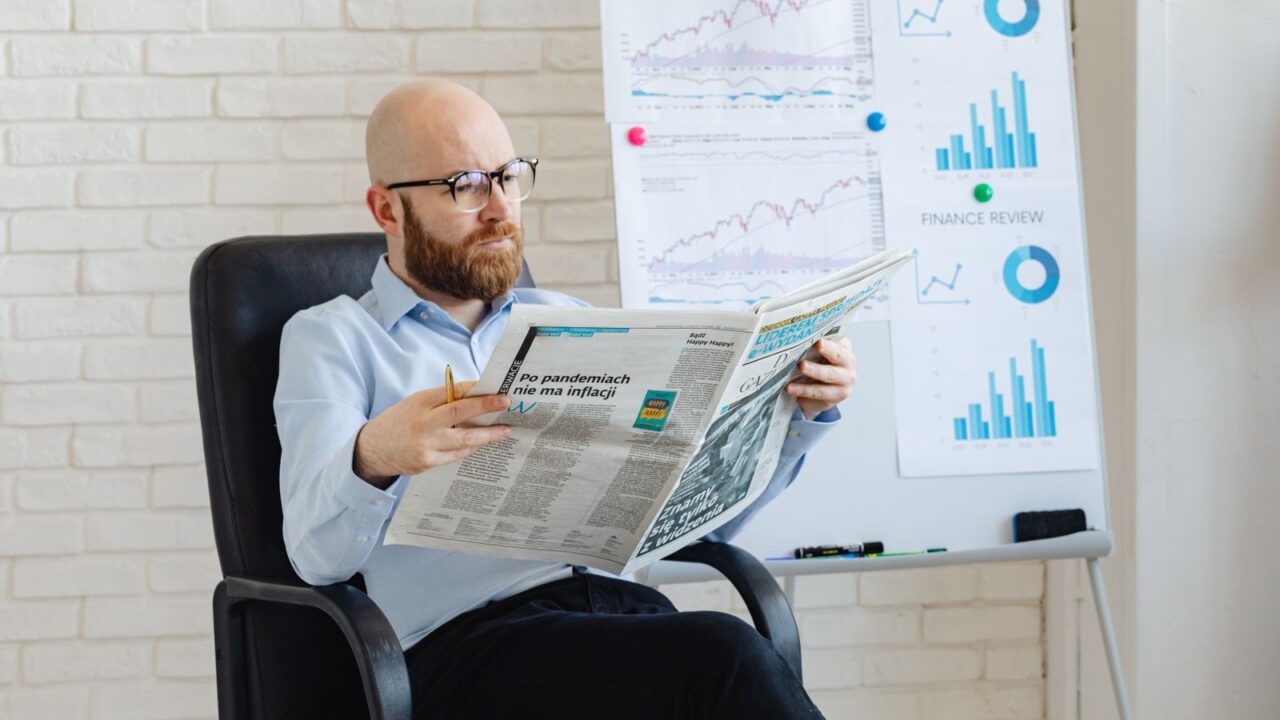 Bald man with beard reading newspaper in office with finance charts on whiteboard.