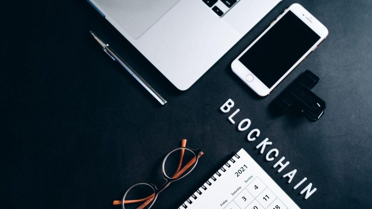 Overhead view of a laptop, smartphone, desk calendar and pen beside the word BLOCKCHAIN.