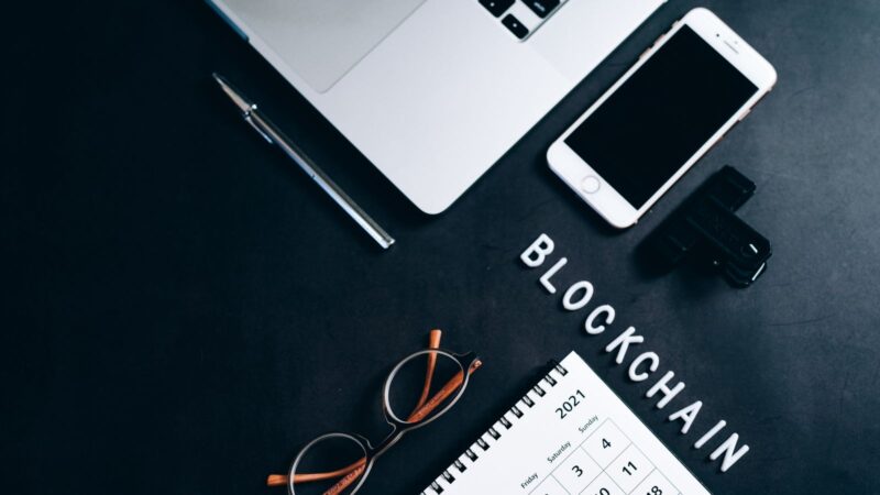 Overhead view of a laptop, smartphone, desk calendar and pen beside the word BLOCKCHAIN.