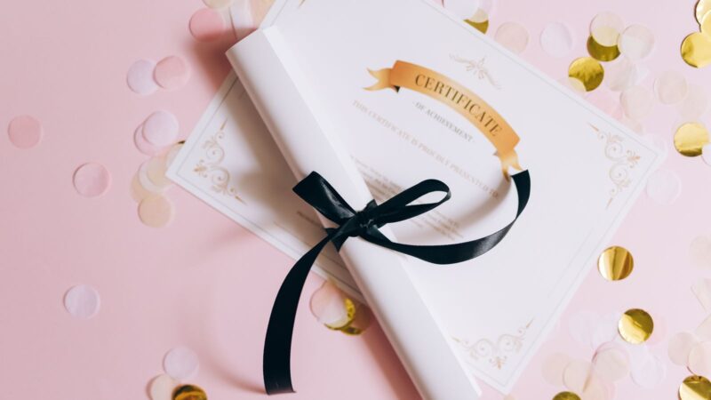 Certificate on a pink surface beside confetti and a rolled document tied with ribbon.